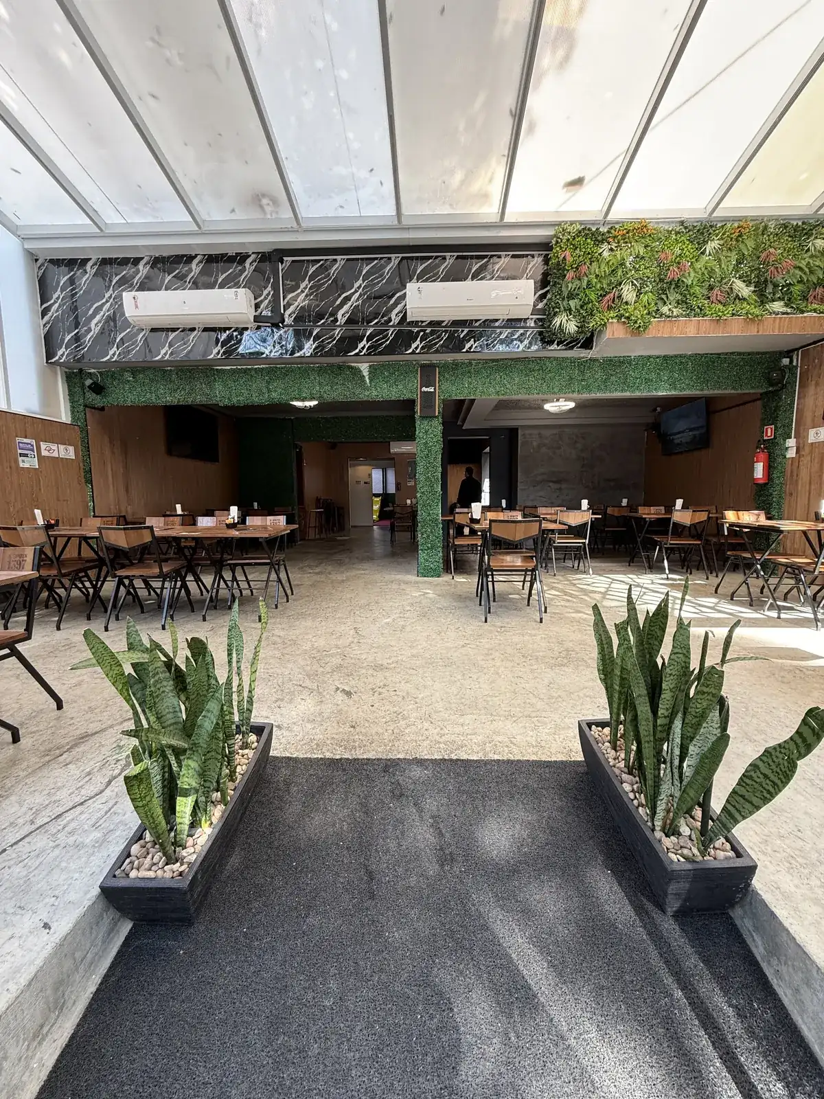 Salão principal do Zora Bar no Gonzaga, com jardim vertical, cobertura translúcida e mesas de madeira